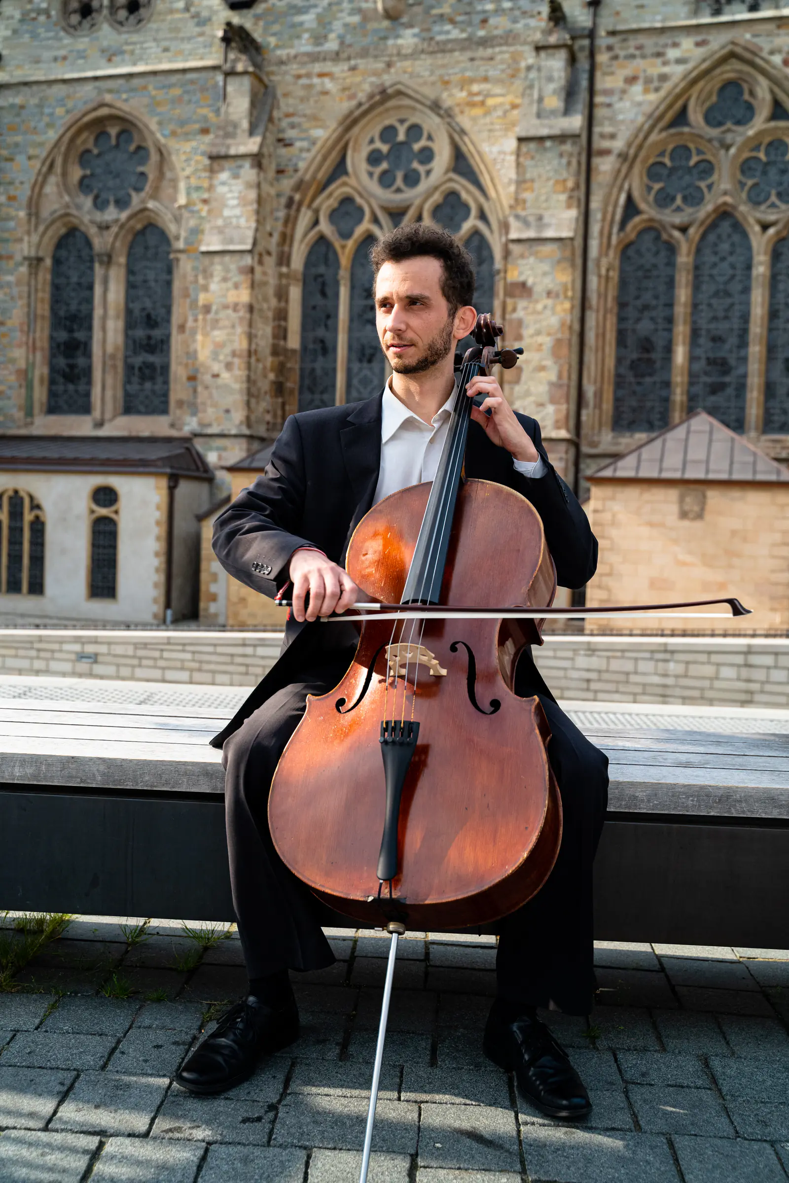 Ventsislav Harkov playing the cello on a bench in front of a Gothic church