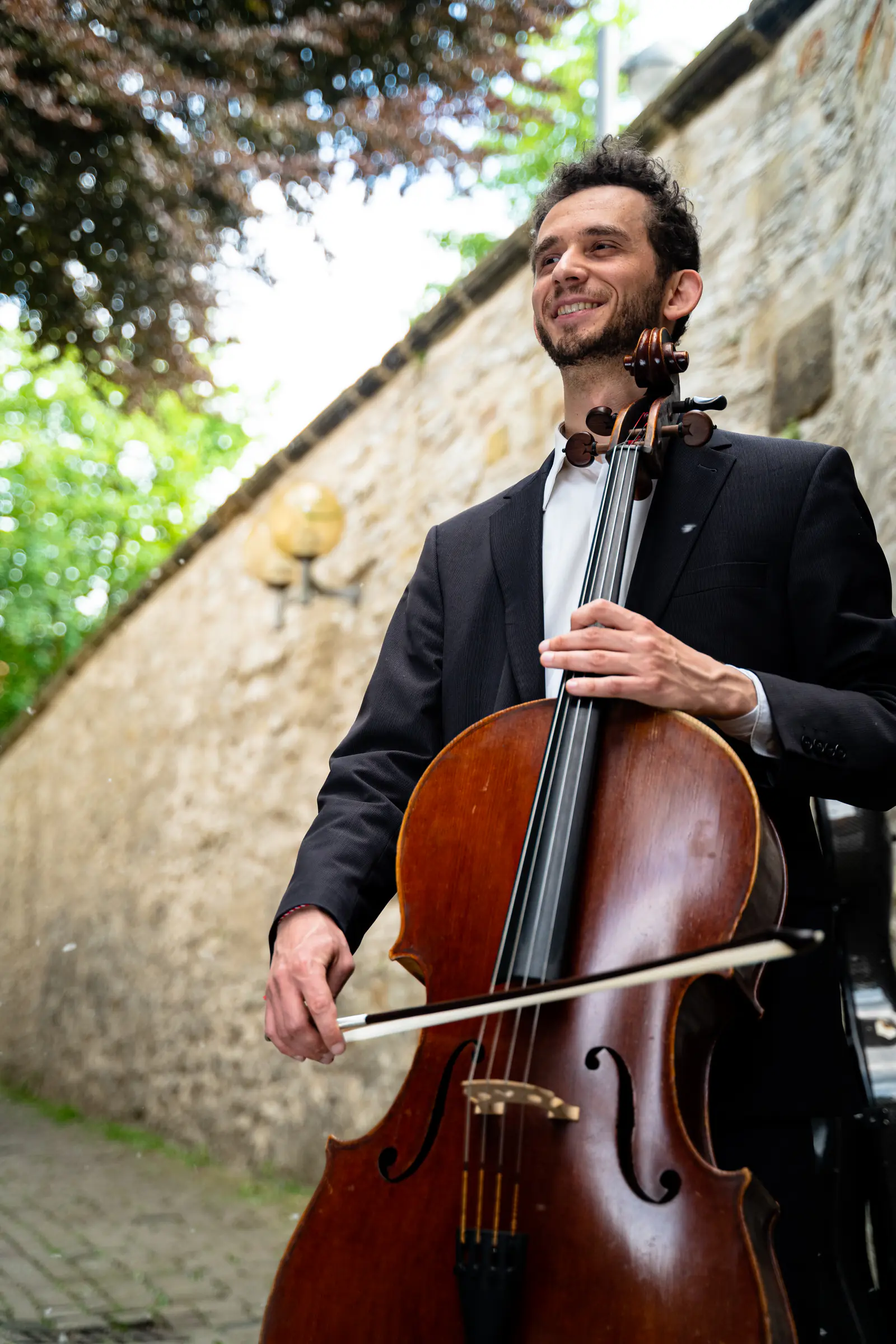 Ventsislav Harkov smiling with his cello and bow beside an old stone wall